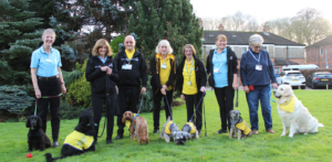 Seven volunteers and their therapy dogs standing on the grass with trees behind them. Dogs are all different breeds.